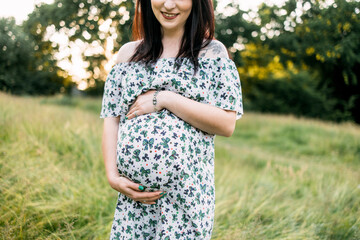 Close up of pregnant caucasian woman in summer dress posing outdoors. Attractive brunette caressing her tummy while standing at green garden.