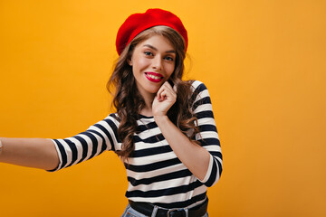 Naklejka premium Optimistic woman in striped outfit smiles and makes selfie. Beautiful lady in good mood with curly hair in red beret looking into camera..