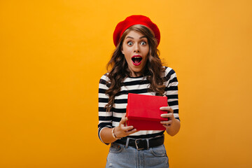 Curly woman looks shocked after opening gift box. Surprised girl in red beret and striped blouse looking into camera rejoices present..