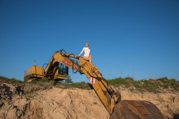 A girl in a white bodysuit in an open space among the sand near an old excavator.