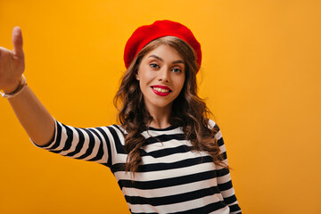 Blue eyed lady in red beret takes selfie on orange background. Curly pretty woman in striped blouse posing on isolated backdrop..