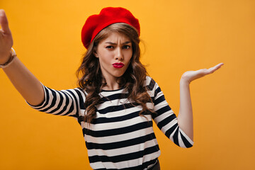 Thoughtful woman shrugs and takes selfie on orange background. Stylish girl with curly hair with red lips in modern clothes posing..