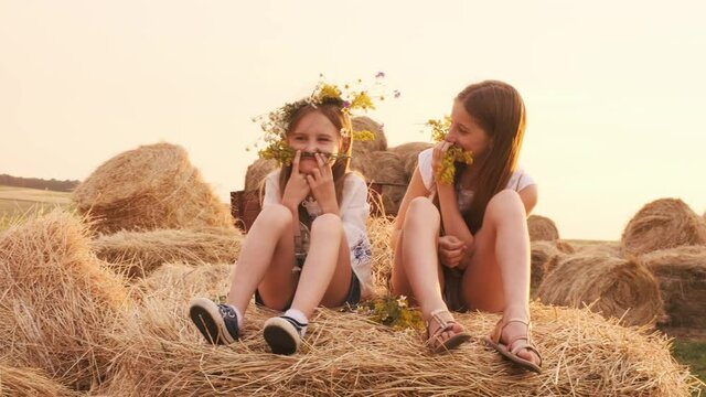 Two pretty sisters wearing wreath sitting on hay stack, having fun and making mustache out of flowers and smiling. Girls spending time together on the nature. Beautiful female teens out of the city