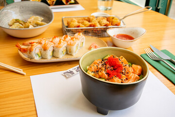 Black bowl with marinated salmon, fish roe, avocado chunks and uramaki rolls with prawns and gyozas on the bottom