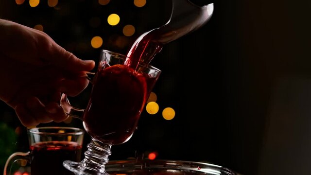 Person hand holding glass mug with orange and pours mulled wine into it indoors with Christmas Eve lights on background. Traditional hot alcohol beverage with spicies at holiday XMas evening