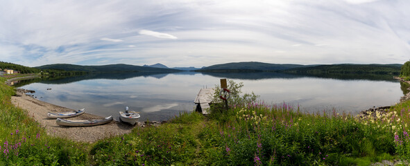 panorama of a calm lake with reflections of mountains and sky and canoes and boat in the foreground