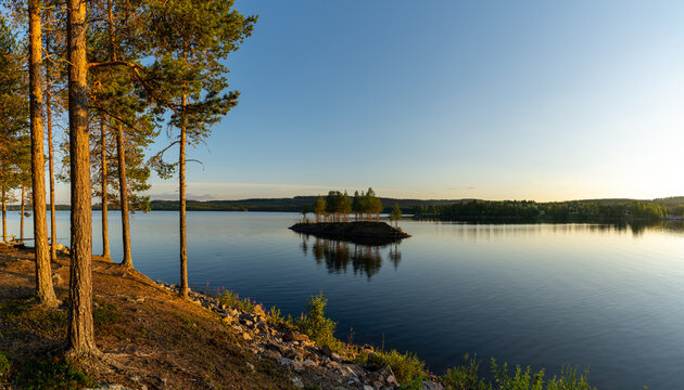 Panorama of a calm lake with small island and golden sunset evening light on the trees and forest on the lakeshore in the foreground
