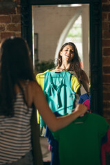 Happy young girl trying on new outfits, preparing for summer vacation standing in front of mirror at home, indoors. Taking right decision. Looks excited, delighted.