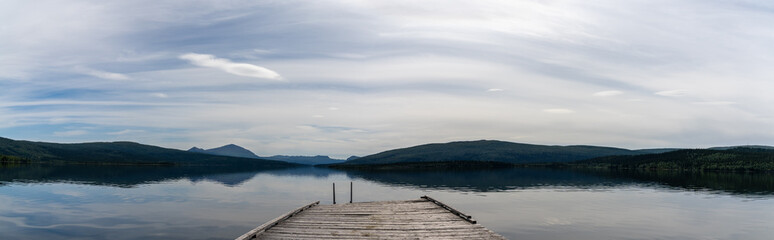 panorama of a calm lake with reflections of mountains and sky and a wooden dock in the foreground
