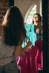 Happy young girl trying on new outfits, preparing for summer vacation standing in front of mirror at home, indoors. Taking right decision. Looks excited, delighted.