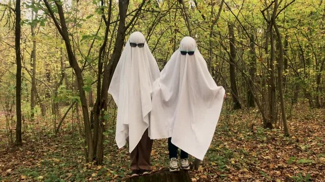 Ghost challenge in the forest or park. Two unrecognizable people teenagers disguised as ghosts with white bed sheets and sunglasses. Jump from a stump to the ground
