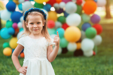 A beautiful, little girl stands against the background of multicolored balloons and shows with her index finger up