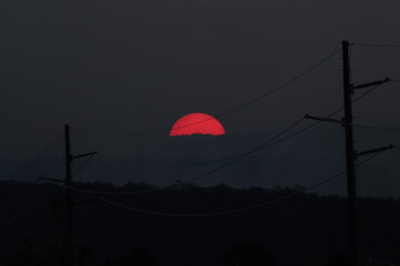 Glowing red sun setting behind the blue mountains with thick bushfire smoke in front and power lines in the foreground.