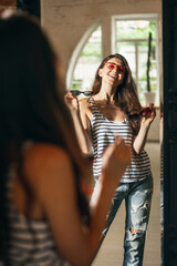 Happy young girl trying on new outfits, preparing for summer vacation standing in front of mirror at home, indoors. Taking right decision. Looks excited, delighted.