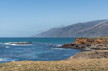 Fototapeta na wymiar San Simeon, CA, USA - June 8, 2021: Pacific Ocean coastline, north of town. Looking at Los Padres Mountain Range at Ragged point south of there. Low red cliffs and blue water under light blue sky.