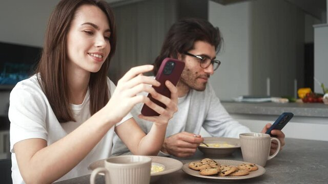 A smiling couple man and woman is using their mobiles while eating breakfast indoors