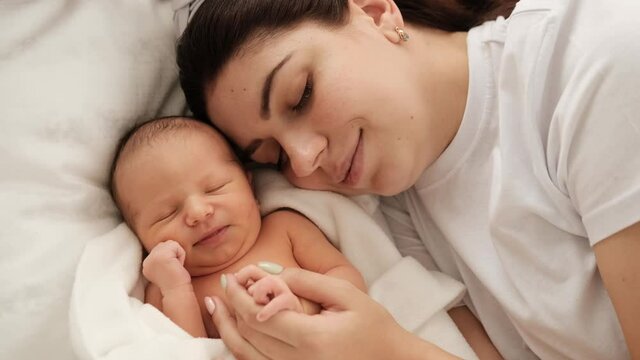 Beautiful young mother lying in the bed with sleeping newborn baby and smiling. Adorable infant child napping with his mom at home. Girl strocking tiny hand of cute resting little kid