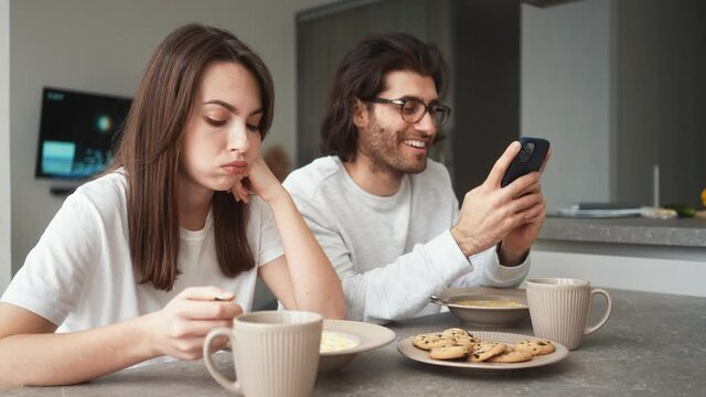 A displeased woman is eating her breakfast while her boyfriend is sitting and using his mobile indoors