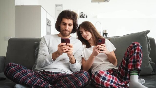A positive couple woman and man are looking social media content using mobiles sitting on the sofa indoors