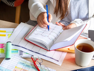 Close-up Of A woman writing schedule In calendar notebook on a desk with paperwork and coffee cup....