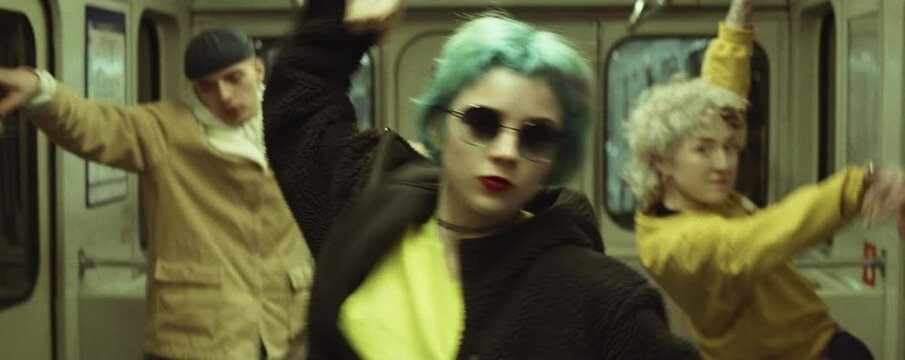 Anamorphic shot of young trendy dressed woman in sunglasses performing expressive modern dance to camera while dancing with team in subway car