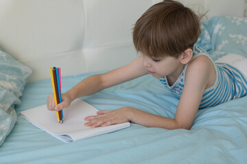 Caucasian boy lies on the bed in the morning and draws with pencils on paper. The student is passionate about the lesson.