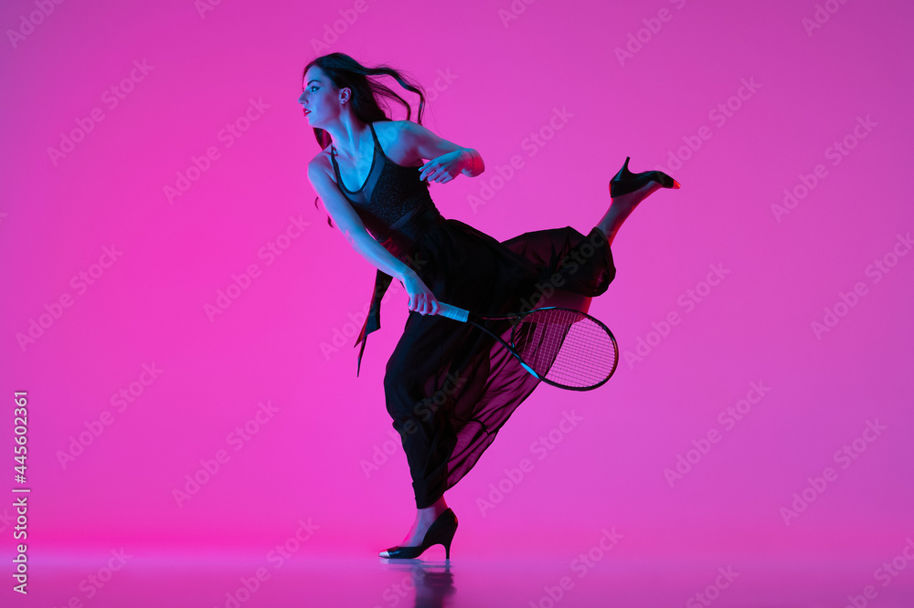 Wall mural High-fashion styled young woman in black evening gown, dress playing tennis isolated over pink neon background. Concept of fashion and sport