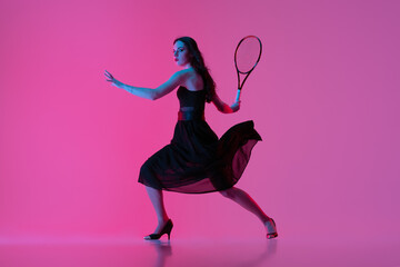 High-fashion styled young woman in black evening gown, dress playing tennis isolated over pink neon background. Concept of fashion and sport