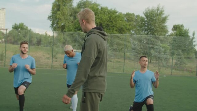 Young football trainer using whistle while team of diverse multiethnic soccer players in sportwear performing hip flexor stretching exercise outdoors at daybreak. Male sportsmen stretching legs
