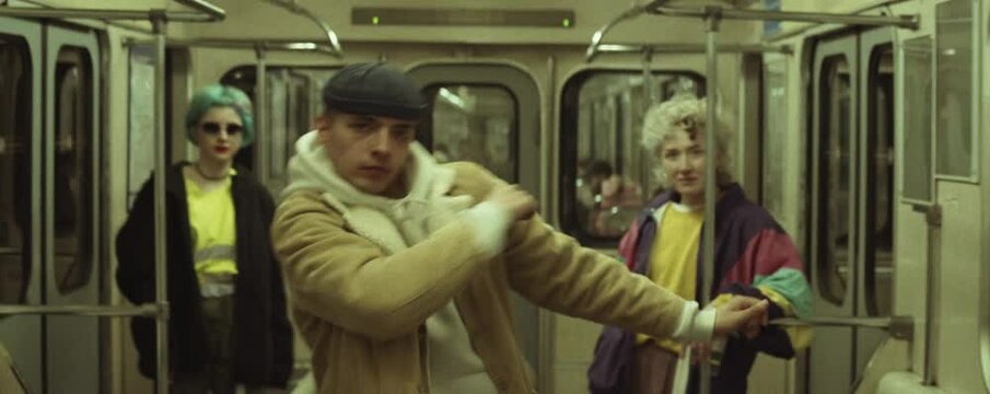 Anamorphic shot of young male dancer in streetwear performing hip hop to camera while dancing with two girls in subway train