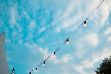 Fashion decoration string lights hanging in restaurant or cafe in the garden at summer party. Outdoor electric lamps. Bright blue sky