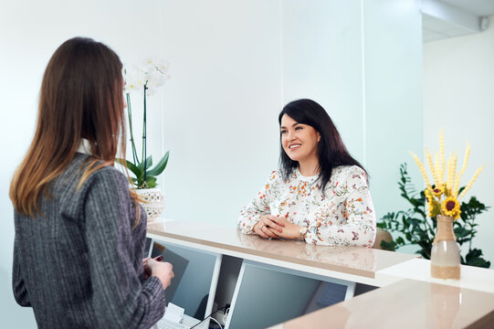 Mid adult woman with beautiful smile talking with young female receptionist, checking appointment in dentist clinic