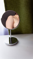 Figure of a fat woman is reflected in a round mirror. Mirror stands on a white table on green curtains background