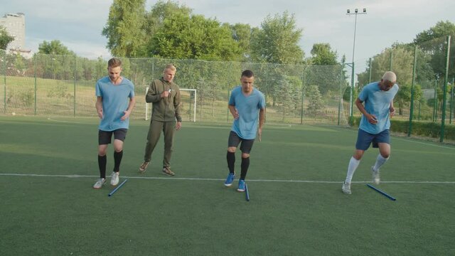 Team of diverse multiracial soccer players in football uniform performing jog on the spot exercise outdoors at sunrise, while young trainer urging sportsmen to move faster, looking on hand watch