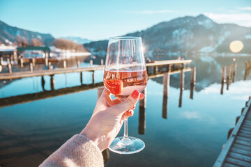 Female hand with glass of rose  wine. Cozy pier on the coast of the lake Tegernsee. Alpine mountains in Bavaria. Mountain view, beautiful landscape in Germany. Adventure in Europe.