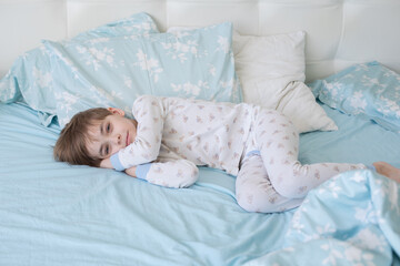 Caucasian boy in pajamas sleeps in the morning on the bed. Top view. The child wakes up with a smile