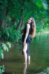 A beautiful young romantic girl with long flowing hair in a black long dress stands in the green water of a summer river or lake.
