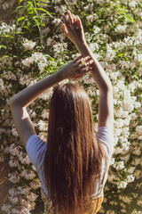 Fototapeta premium Beautiful slender brunette girl raised her hands up and posing back against the background of flowering bushes on a sunny day
