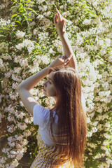 Beautiful slender brunette girl raised her hands up and posing against the background of flowering bushes on a sunny day