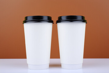 Close up of two white cardboard cup of coffees on white table against brown background. Concept of hot drinks for take away