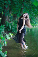 A beautiful young romantic girl with long flowing hair in a black long dress stands in the green water of a summer river or lake.A beautiful young romantic girl with long flowing hair in a black long 