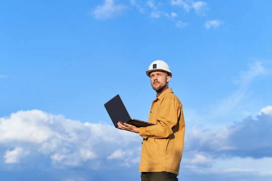 Beautiful caucasian bearded building contractor, foreman or engineer in white helmet and orange jacket using laptop at construction site at sunny day with blue cloudy sky. High quality image