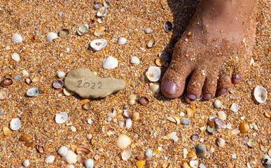 The numbers 2022 are on a small gray stone against the background of a small shell next to a woman's leg. The concept of the New Year 2022. Summer holidays and sea trips.