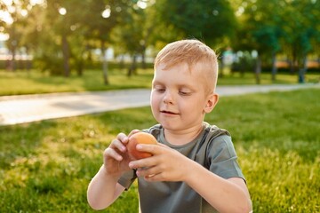 A small five-year-old red-haired boy looks at a peach and is about to eat it in the park, selective focus on the face