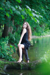 A beautiful young romantic girl with long flowing hair in a black long dress stands in the green water of a summer river or lake.