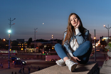 Fototapeta premium Beautiful young girl sits on evening city background. Stylish student girl lonely sitting on the high step