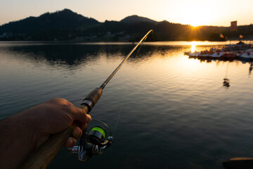 Hand holding fishing rod and a beautiful sunrise over calm lake with mountains in the distance. Colibita lake, Romania