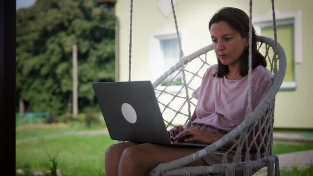 Portrait Of Woman Working Outdoors In Her Garden. Young Woman Using Laptop Computer While Sitting In Cozy Garden Swing Chair At Backyard Of Her House