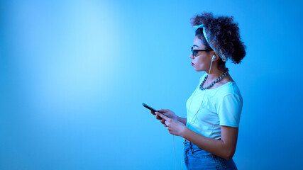 Trendy girl with afro curls uses a smartphone and earphones on a blue studio background with copy space, online conversation, listens to music