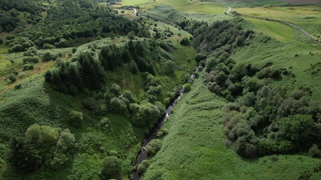 Aerial footage over a river flowing through a partially wooded glen with a long and winding rural road disappearing into the distance.